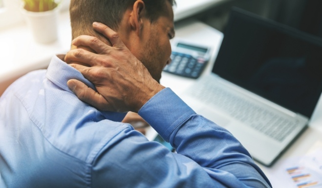 Man sitting at desk and wincing while holding the back of his neck